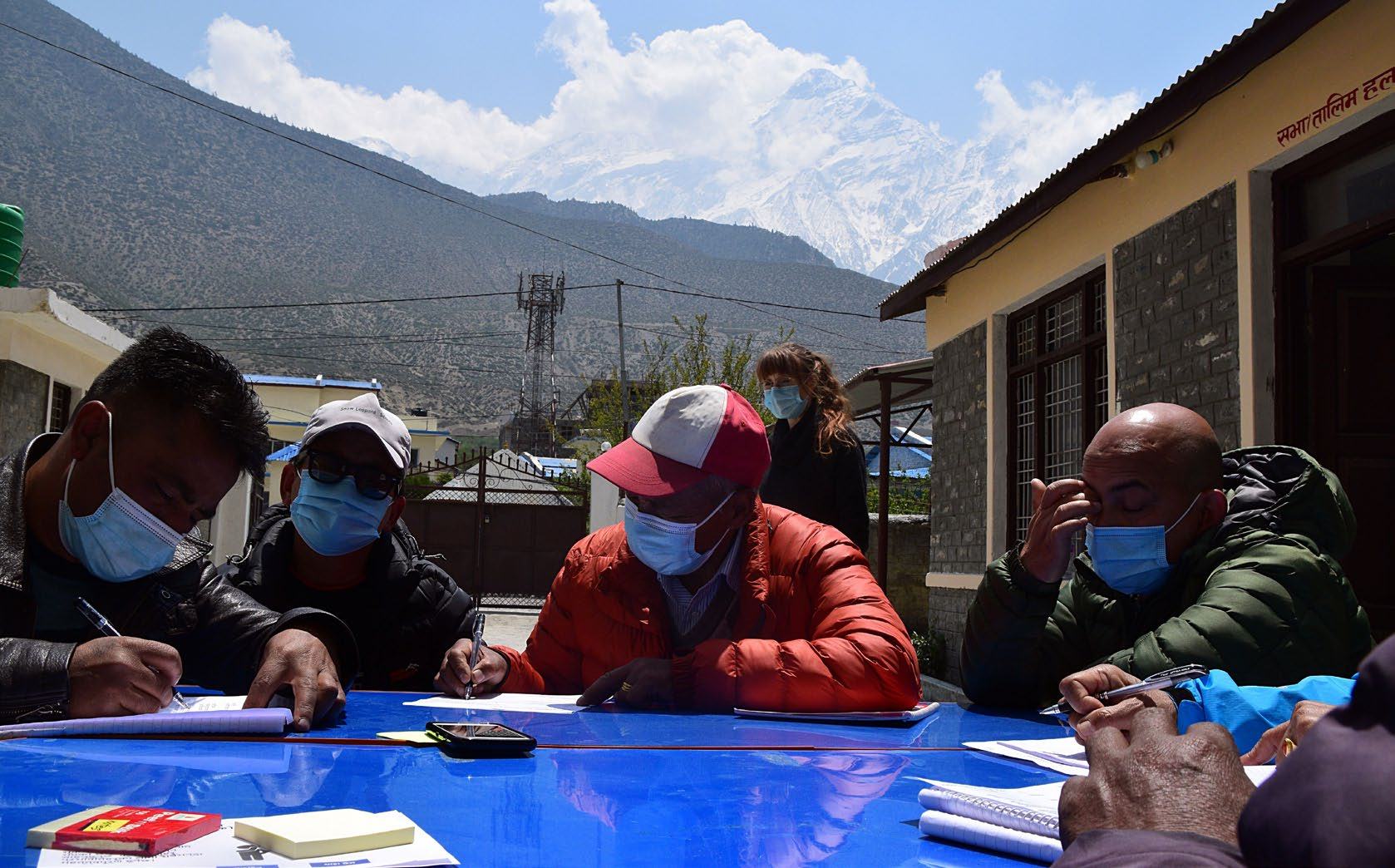 Several men in masks writing at a blue outdoor table, with a snowy mountain landscape in the background, overseen by a woman.