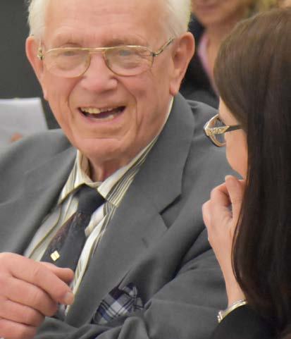 A smiling elderly man in glasses and a suit talking with a woman, possibly at a meeting or social event.