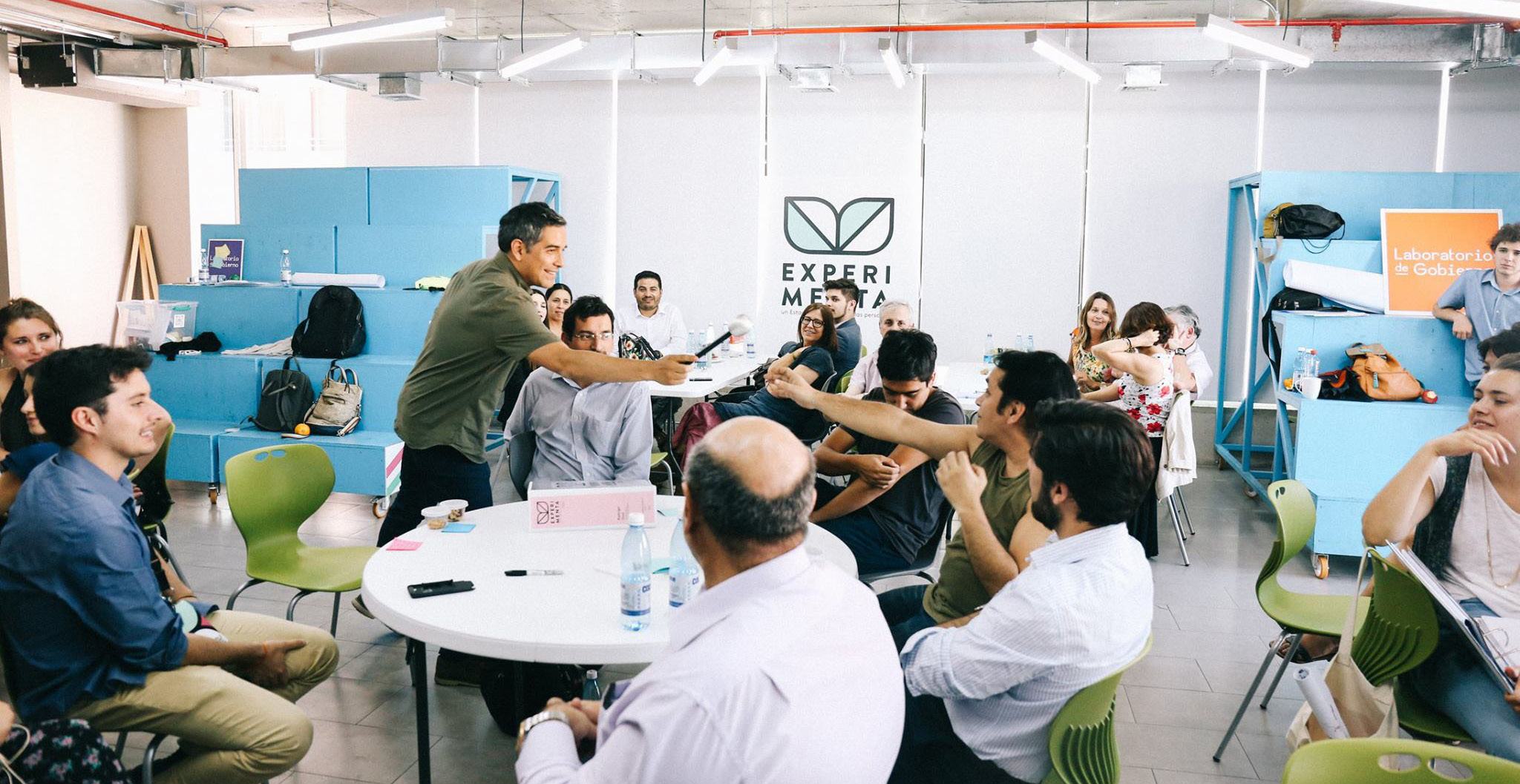 Man with a microphone engaging with a diverse group of people seated at tables during a presentation or workshop.