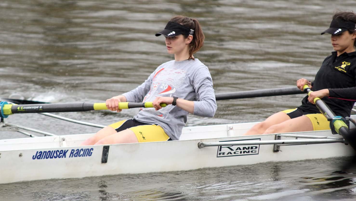 Two women in a Janousek Racing boat, actively rowing with oars on a body of water.