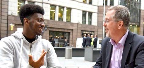 Two men of different ages smile and talk while sitting outdoors, with city buildings visible in the background.