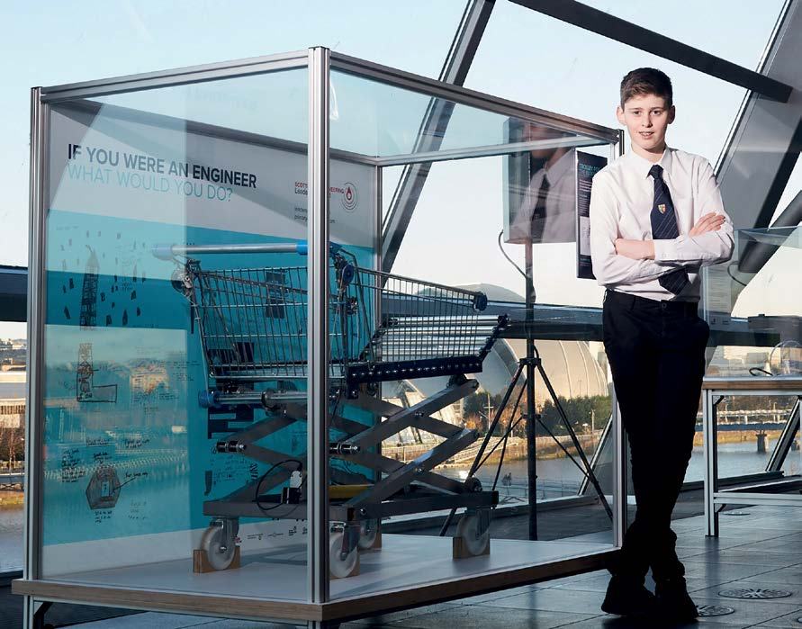 A young man stands next to a glass display case containing a modified shopping cart, part of an engineering exhibit.