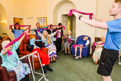 An instructor leading a group of elderly residents in an exercise class using pink resistance bands in a brightly lit common room.