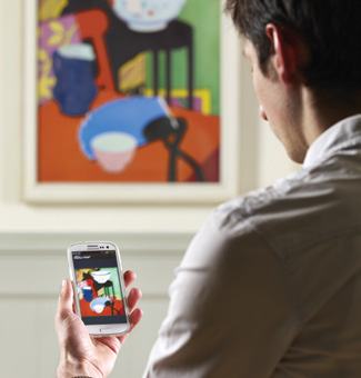 A person holding a smartphone displaying a digital image of a painting, while the actual painting hangs on the wall in the background.