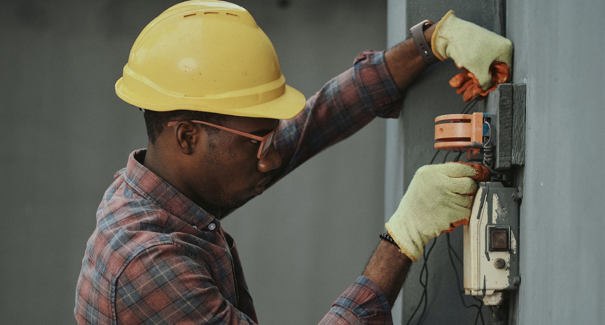 Construction worker in a yellow hard hat and gloves performing electrical wiring or installation work on a wall.