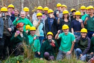 A group of people in yellow hard hats and green shirts holding mugs outdoors in a wooded area.
