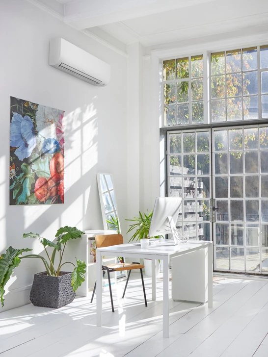 A white Daikin indoor air conditioning unit installed high on the wall of a bright, modern office with large windows.