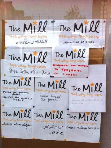 A collage of signs on a board, all featuring "The Mill" and "People making things happen" translated into multiple languages, showcasing community outreach.