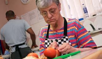An older woman in glasses and an apron peeling tomatoes at a counter, with a man in the background.