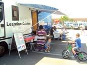 Mobile Learning Centre truck parked outdoors with people engaging at tables, and a child riding a bicycle in the foreground.