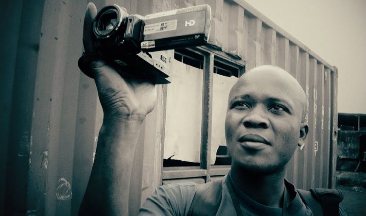 A man holding a video camera, recording something, against an industrial container background.