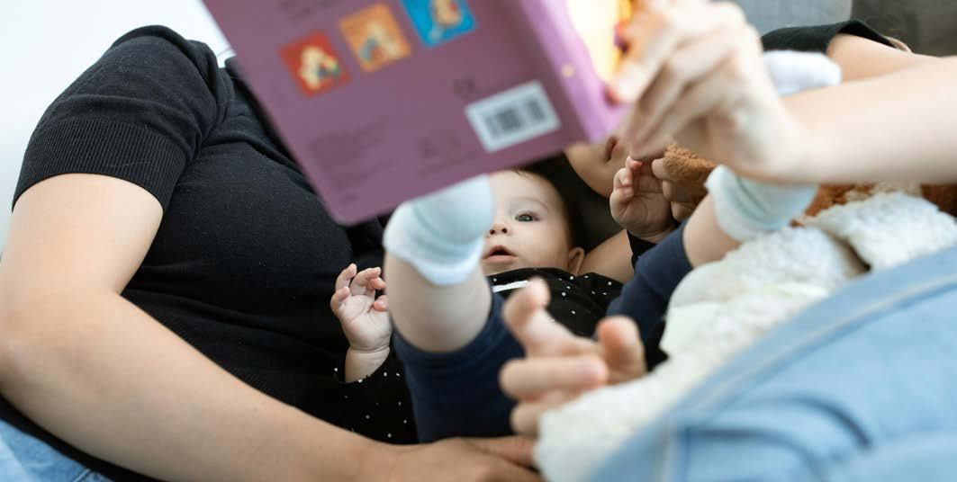Close-up photo of an adult's arm holding an open book for a baby lying down and looking up at the pages.