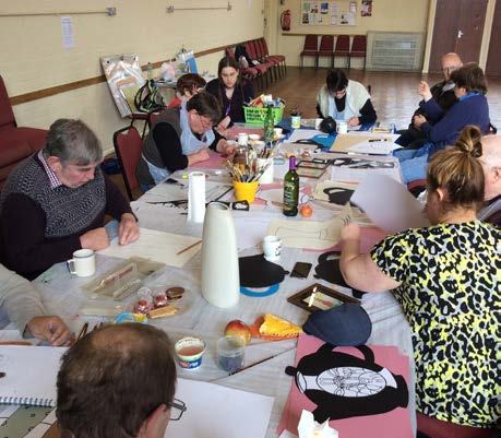 Older adults participate in a craft activity around a long table, with various art supplies and paper cutouts.