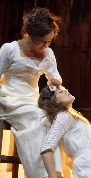 Two women in white dresses on a stage. One sits, gently touching the head of the other who is leaning back, eyes closed.