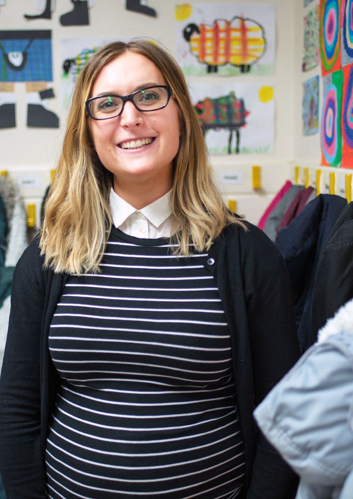 Smiling woman with blonde hair and glasses, wearing a striped shirt, standing in a room with children's artwork.