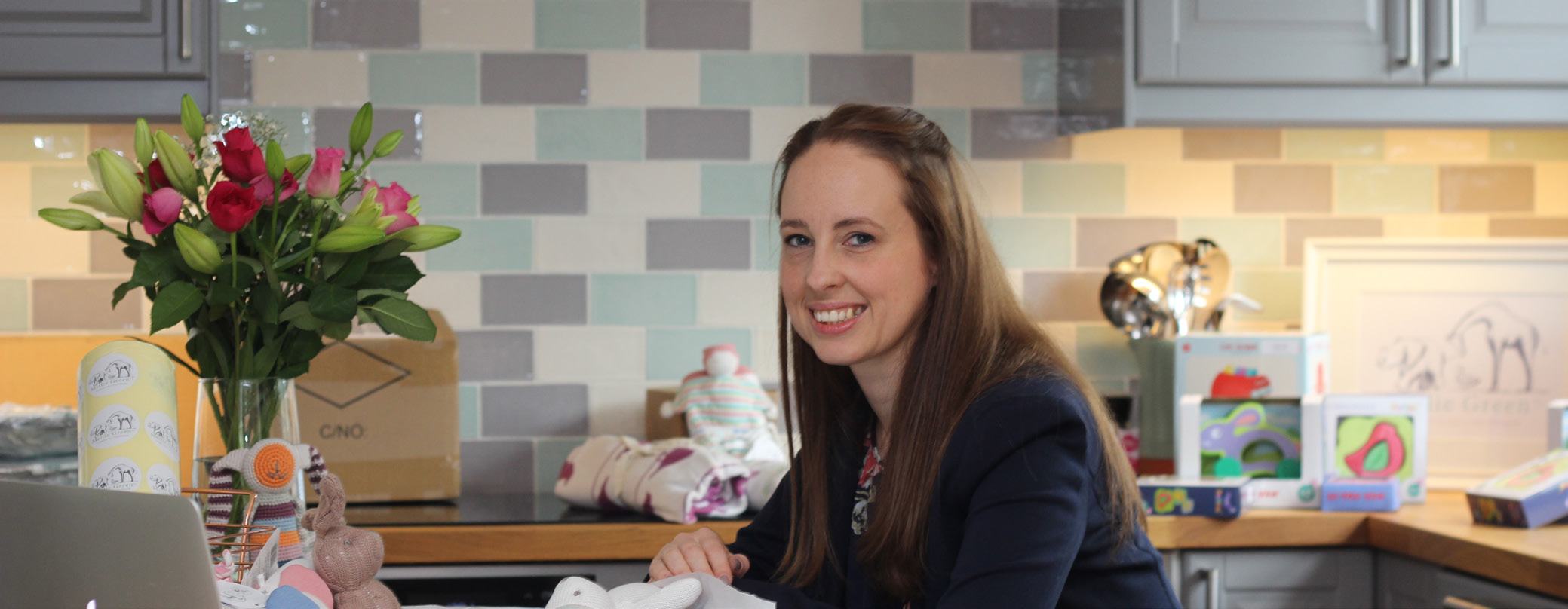 Headshot of a smiling woman seated at a kitchen counter with flowers, boxes, and toys, suggesting a home business or creative work.