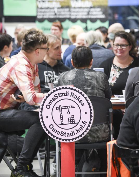 People at a workshop, with a foreground sign reading "OmaStadi Raksa OmaStadi.hel.fi" featuring a crane and buildings icon.