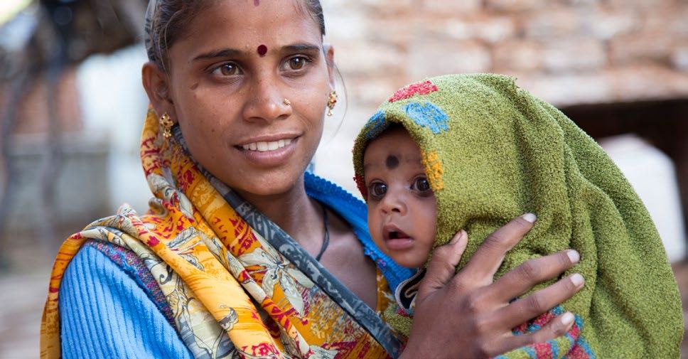 A smiling woman with a bindi holds a baby wrapped in a green blanket, both looking towards the viewer.