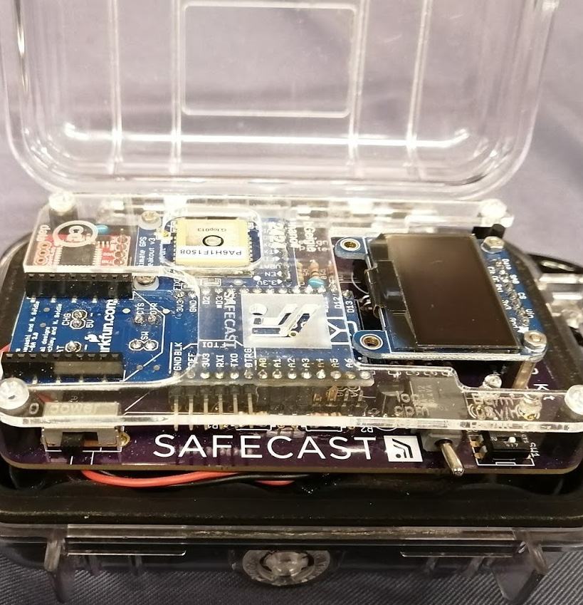 Close-up of a Safecast radiation sensor, showing its circuit board, display, and components housed in a transparent case.