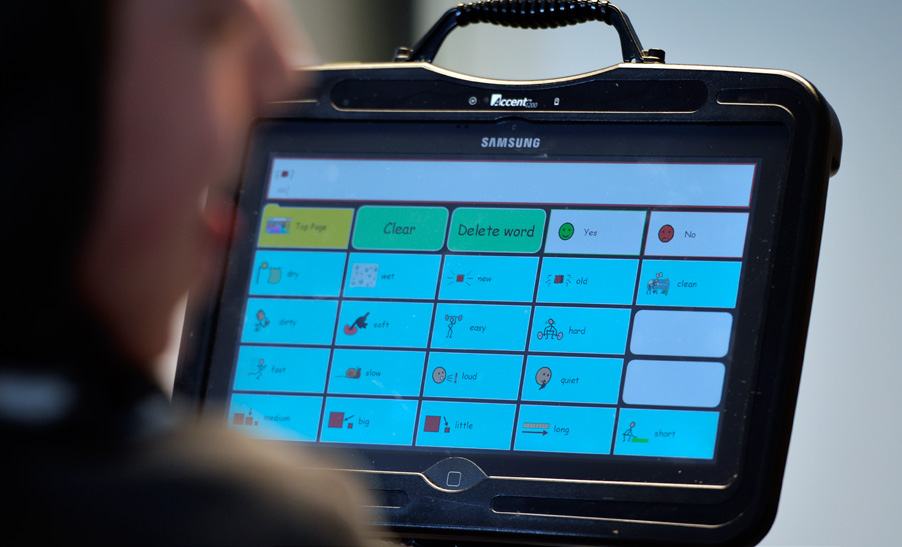 A person interacting with an AAC tablet displaying buttons for words like 'dry', 'wet', 'new', 'old', 'clean', 'dirty', 'soft', 'easy', 'hard', 'fast', 'slow', 'loud', 'quiet', 'medium', 'big', 'little', 'long', 'short'.