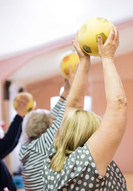 Group of older adults from behind, raising yellow exercise balls above their heads during a fitness class.