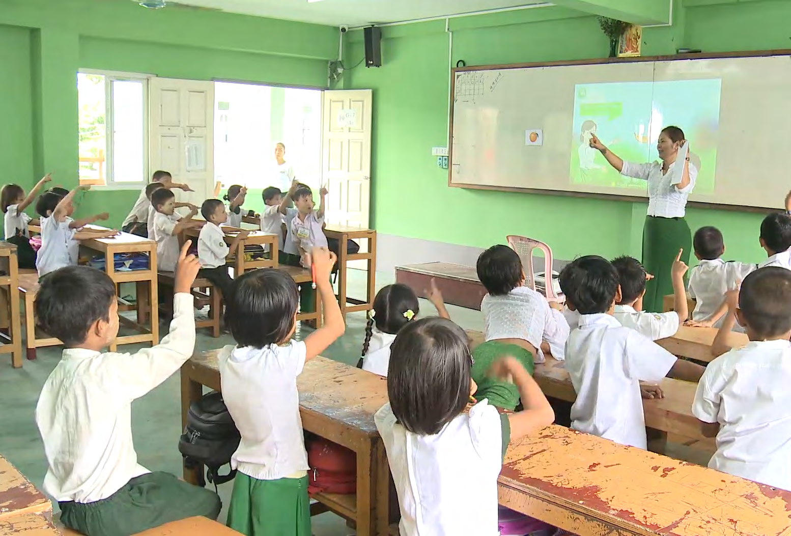 Teacher instructs young students in a classroom using a projector, with many students raising their hands, indicating active participation.