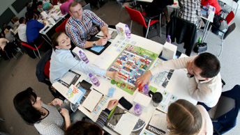 Group of people collaborating around a table with a board game, likely a workshop or training simulation.