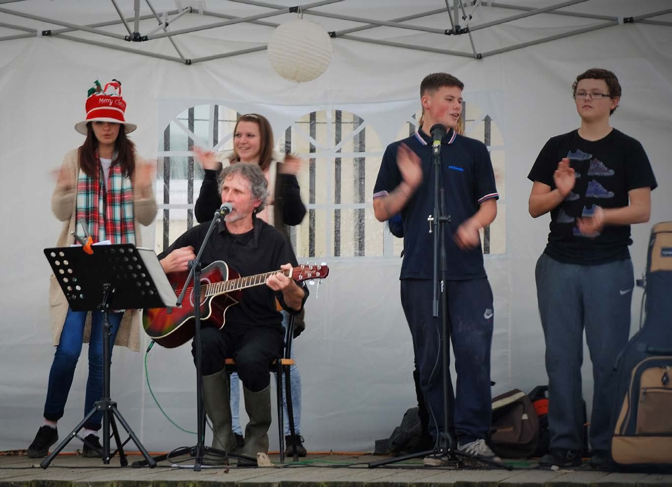A man playing an acoustic guitar and singing under a tent, with several other people around him, some clapping.