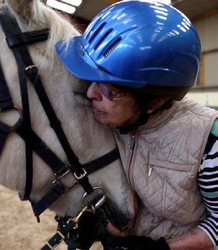 A woman in a blue helmet and vest gently kisses a white horse's face, showing affection.