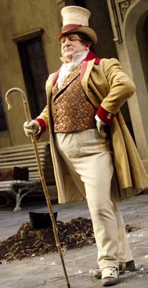 A man in a period costume with a top hat, long coat, and waistcoat, holding a walking stick, stands on a stage set.