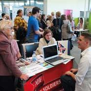 An indoor event scene with multiple people, one booth displaying a laptop and materials for "ABF The Soldiers' Charity" as people interact.
