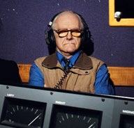 An older man with headphones and glasses, looking intently, with audio equipment in front of him, suggesting a studio setting.