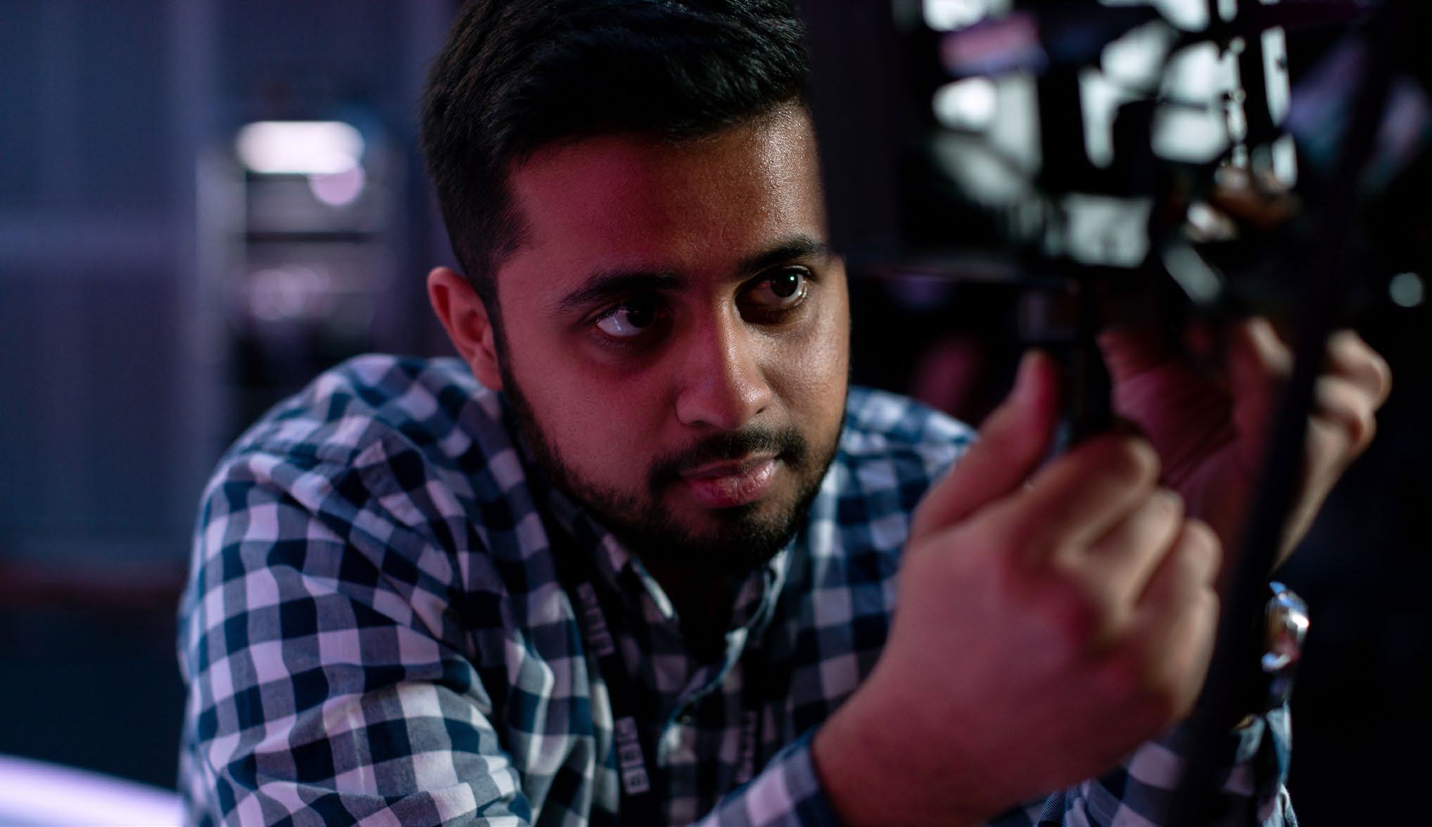 A man in a plaid shirt intently adjusting technical equipment, possibly a camera, in a professional setting.
