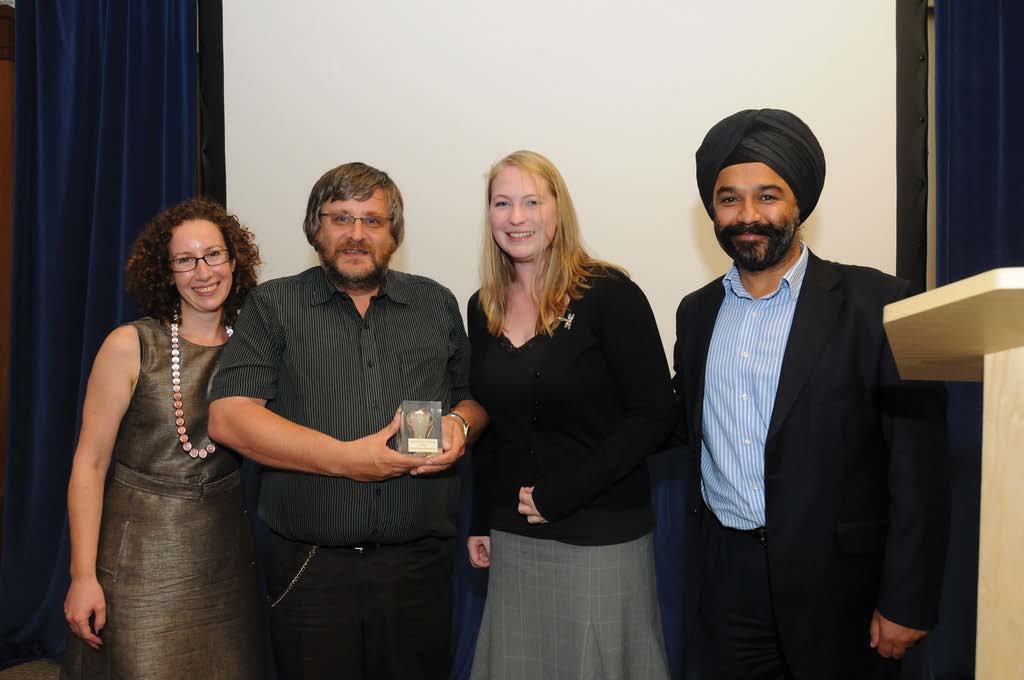 Four people stand together; a man in the center holds an award, likely at an event with a podium and screen in the background.