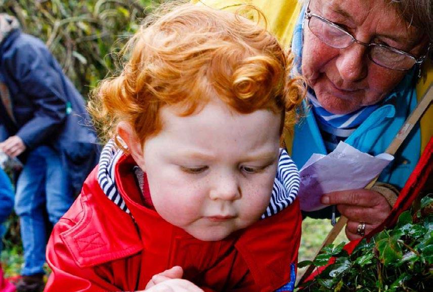A young child with red curly hair in a red raincoat looks intently at something on the ground, with an older adult looking on.