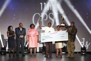 Six people on a stage during an awards ceremony, holding trophies and a large ceremonial check for 'R100 000.00'.