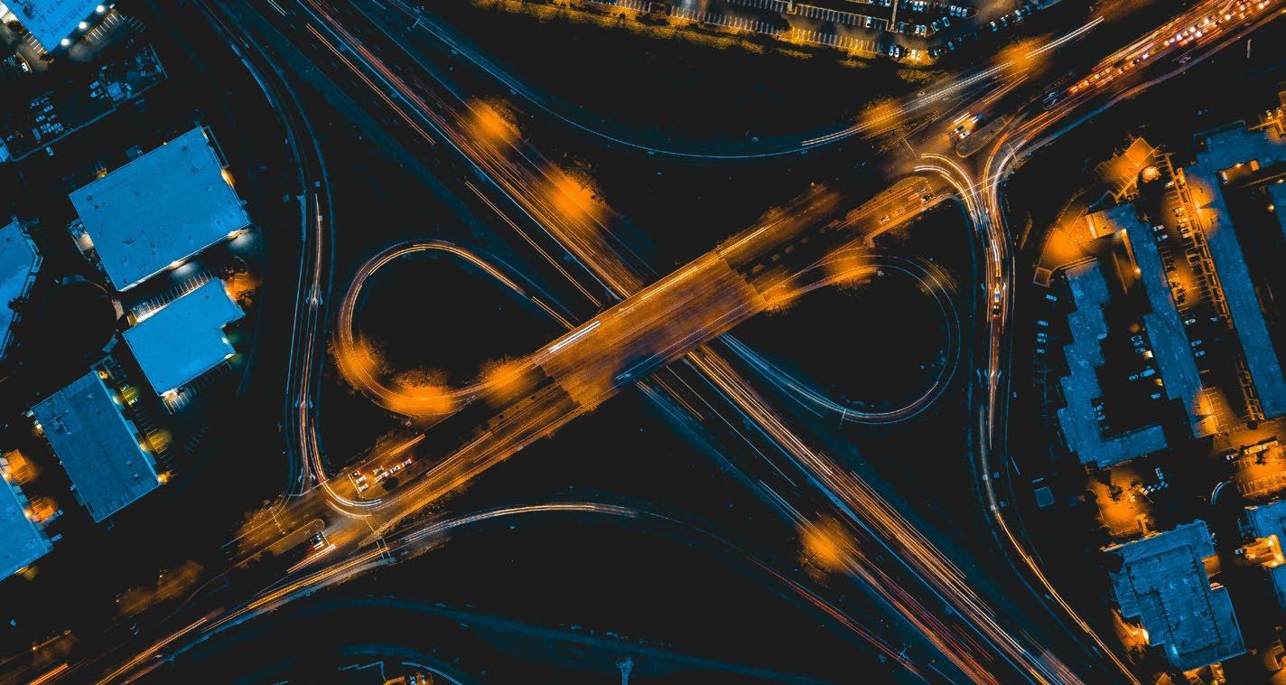 Aerial long-exposure shot of a complex highway interchange at night, showing vehicle light trails and surrounding buildings.