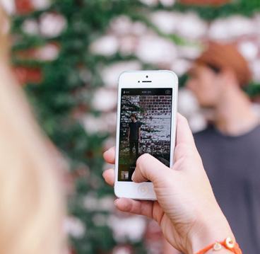 A person holding a smartphone to take a photo of another person against a brick wall.