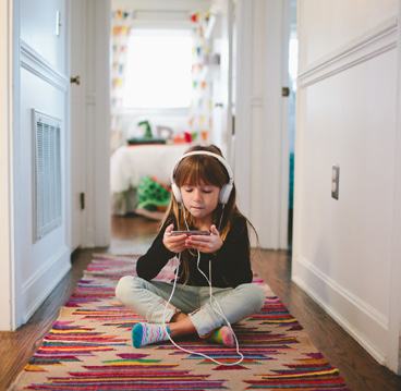 A young girl wearing headphones and looking at a smartphone while sitting on a colorful rug in a hallway.
