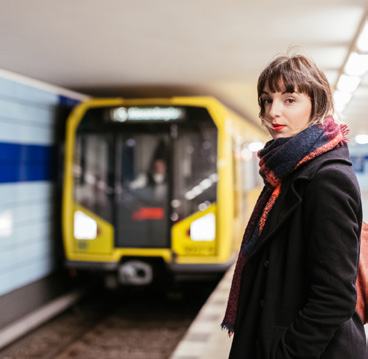 A woman with a red scarf stands on a subway platform, with a yellow train arriving in the background.