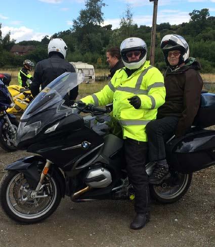 A group of motorcyclists gathered, with a man in a yellow reflective jacket on a black BMW bike with a passenger.