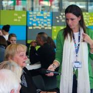 A woman in a green cardigan holding a microphone, speaking to an older seated woman in an indoor setting with colorful wall panels covered in sticky notes.