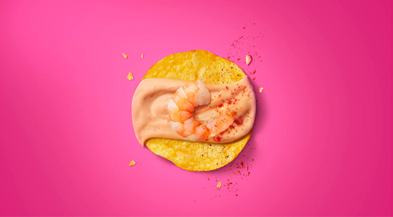 A single tortilla chip topped with shrimp, sauce, and seasoning, on a vibrant pink background.