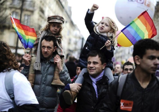 Two men carrying young children on their shoulders, all participating in a parade, with rainbow flags visible.