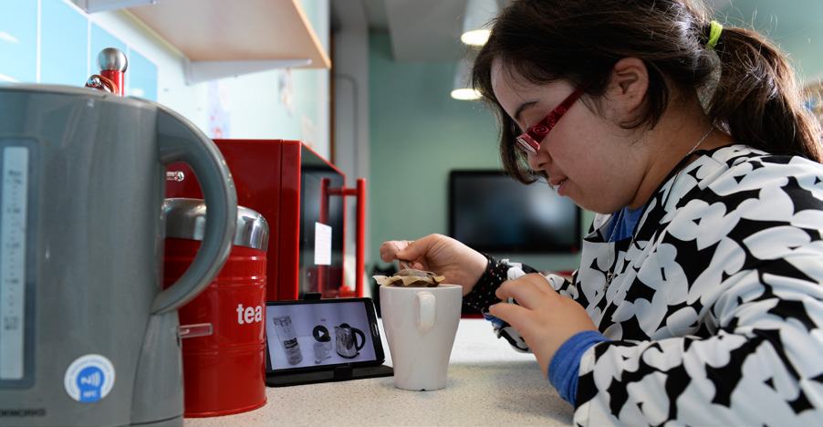 A person following tea-making instructions displayed on a smartphone while preparing a mug of tea in a kitchen, with a kettle nearby.