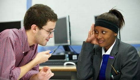 Male teacher talking to a female student in a classroom setting, with computers in the background.