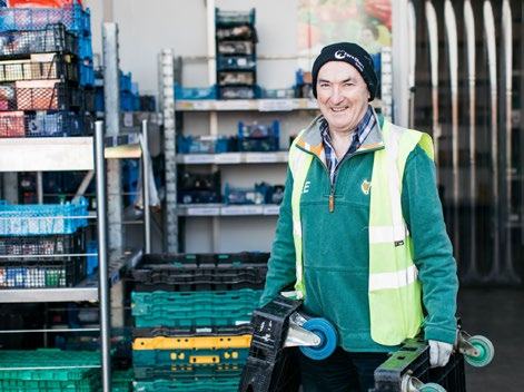 A smiling man in a high-vis vest and beanie holds a hand truck in a storage area filled with crates of fresh produce.