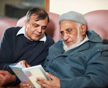 Two men, an older man with a white beard and a younger man, sit side-by-side, engaged in reading a book together.