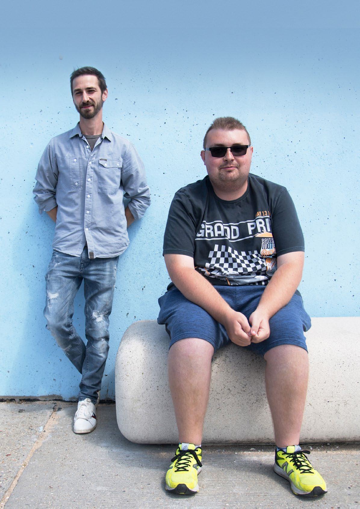 Two men, one leaning against a blue wall and another sitting on a concrete bench, looking at the camera.