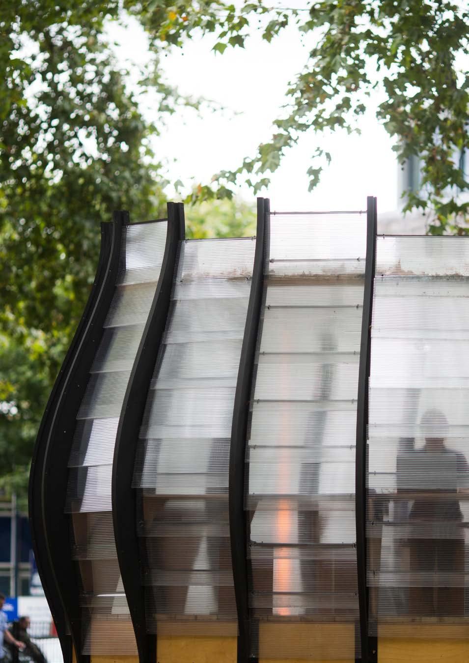 Close-up of a modern architectural structure with translucent, ribbed panels and dark frames, seen through trees.